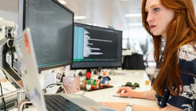 A focused female software engineer coding on dual monitors in a modern office.